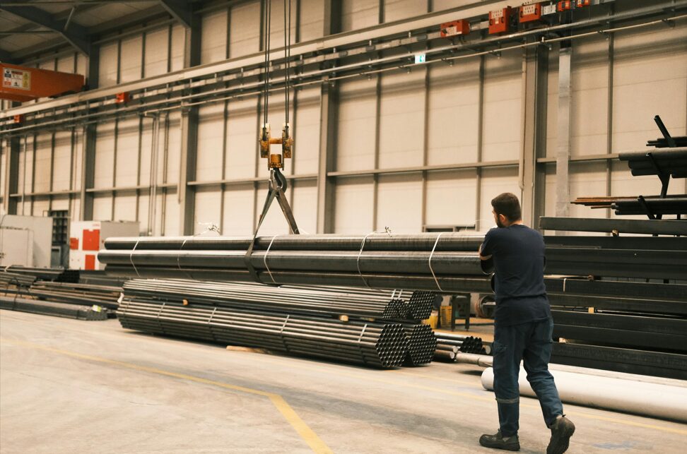 Worker overseeing steel pipe lifting with crane inside industrial warehouse